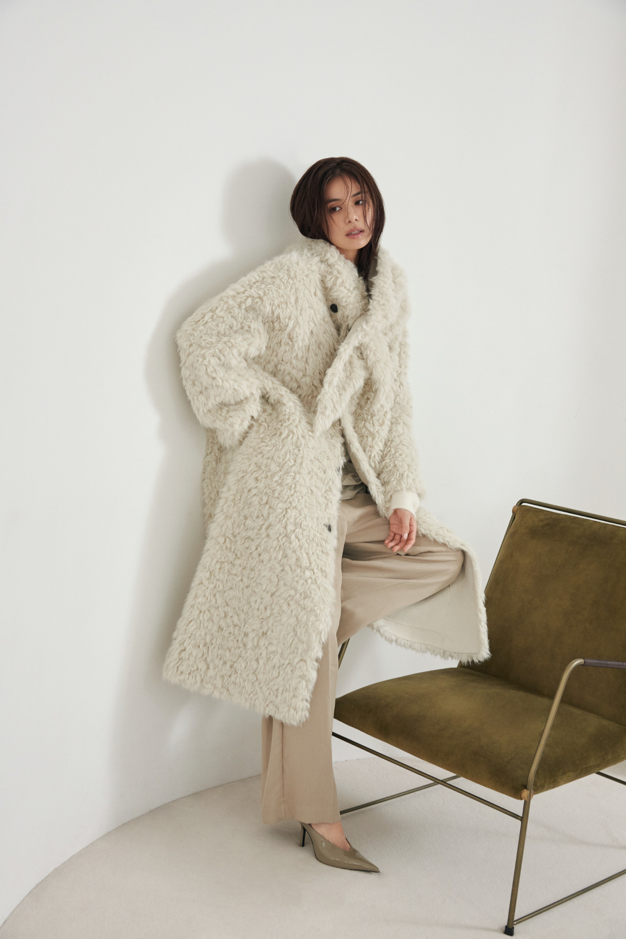Look01 ファーのロングコートとベージュのパンツで椅子の横に立つ着用カット