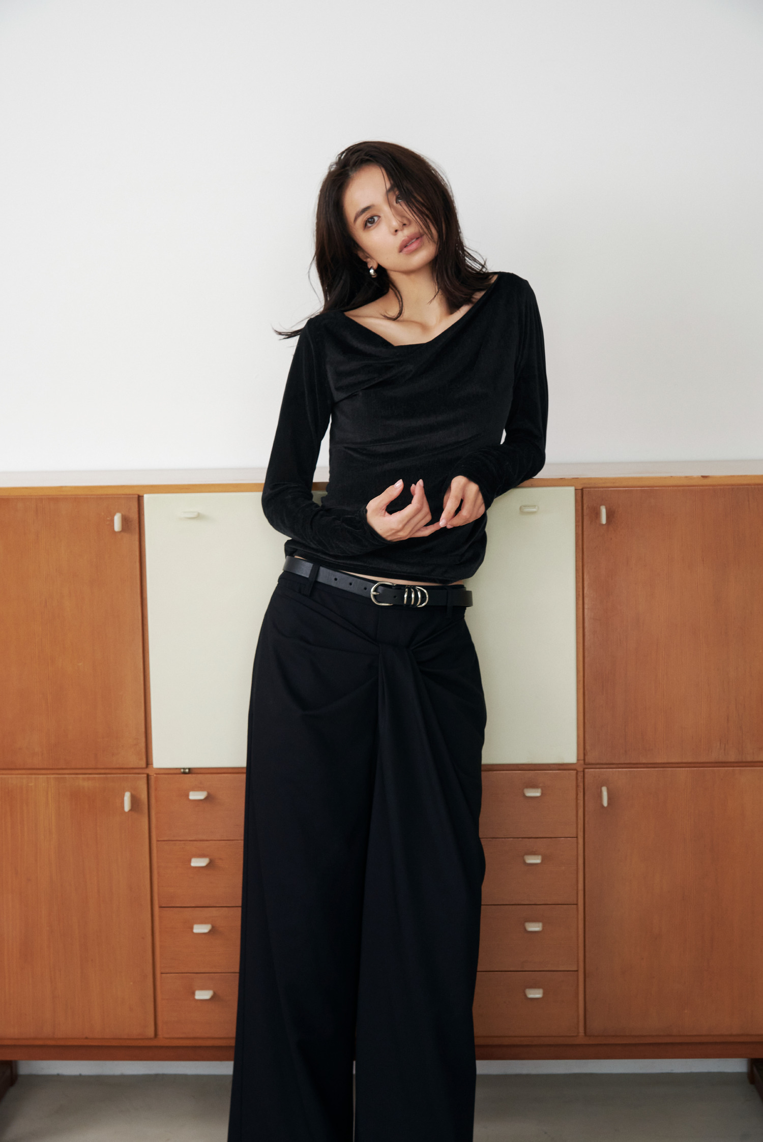 Look11 黒のトップスとパンツで木製キャビネットの前に立つ着用カット