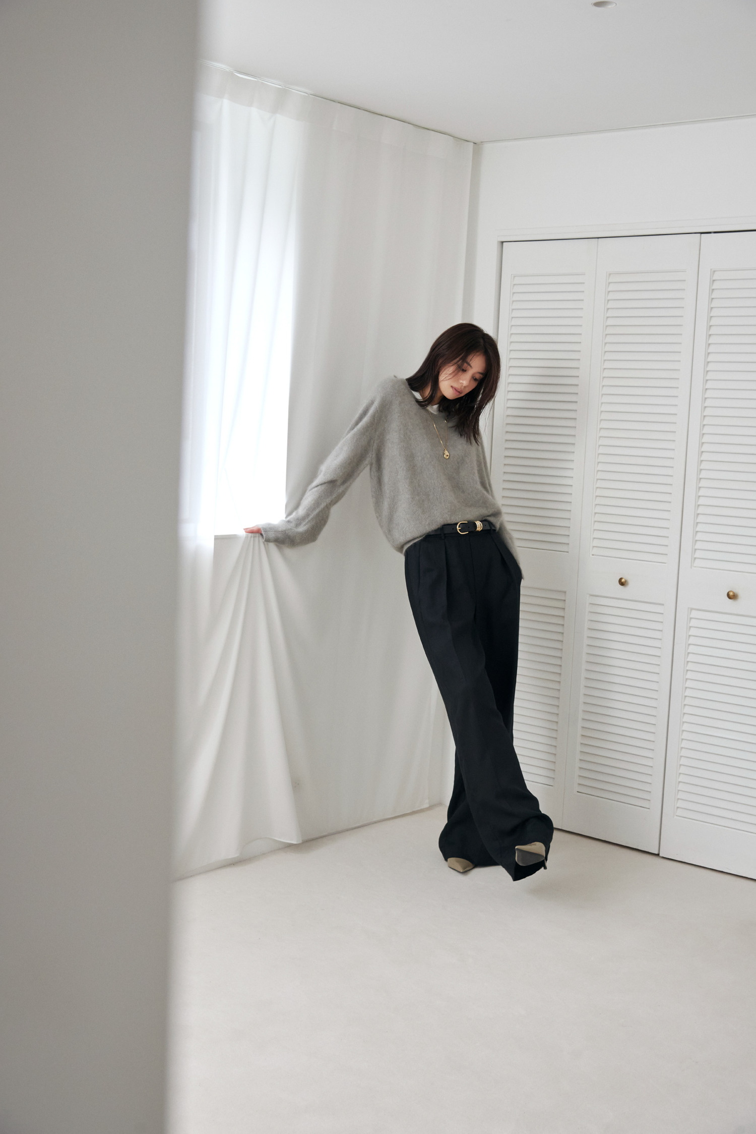 Look12 グレーのニットとカーテンを手に窓辺に立つ着用カット